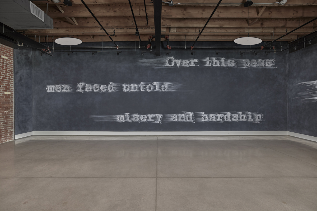 Installation view of Gary Simmons: Rush at Cookie Factory in Denver, showing a chalkboard wall drawing that reads "Over this pass men faced untold misery and hardship."