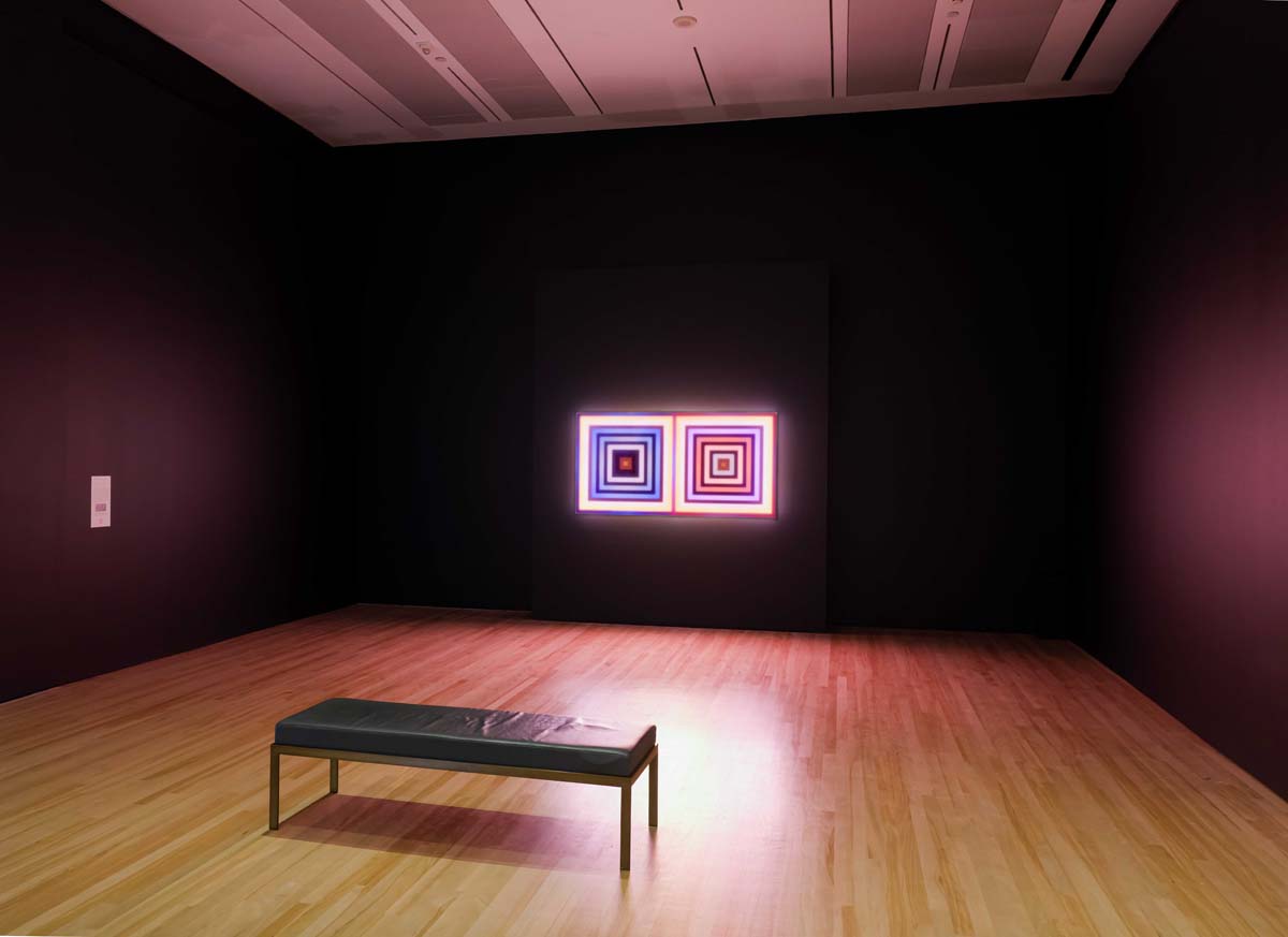 Installation view of Run the Code, with two illuminated squares at the end of a dark room with a bench