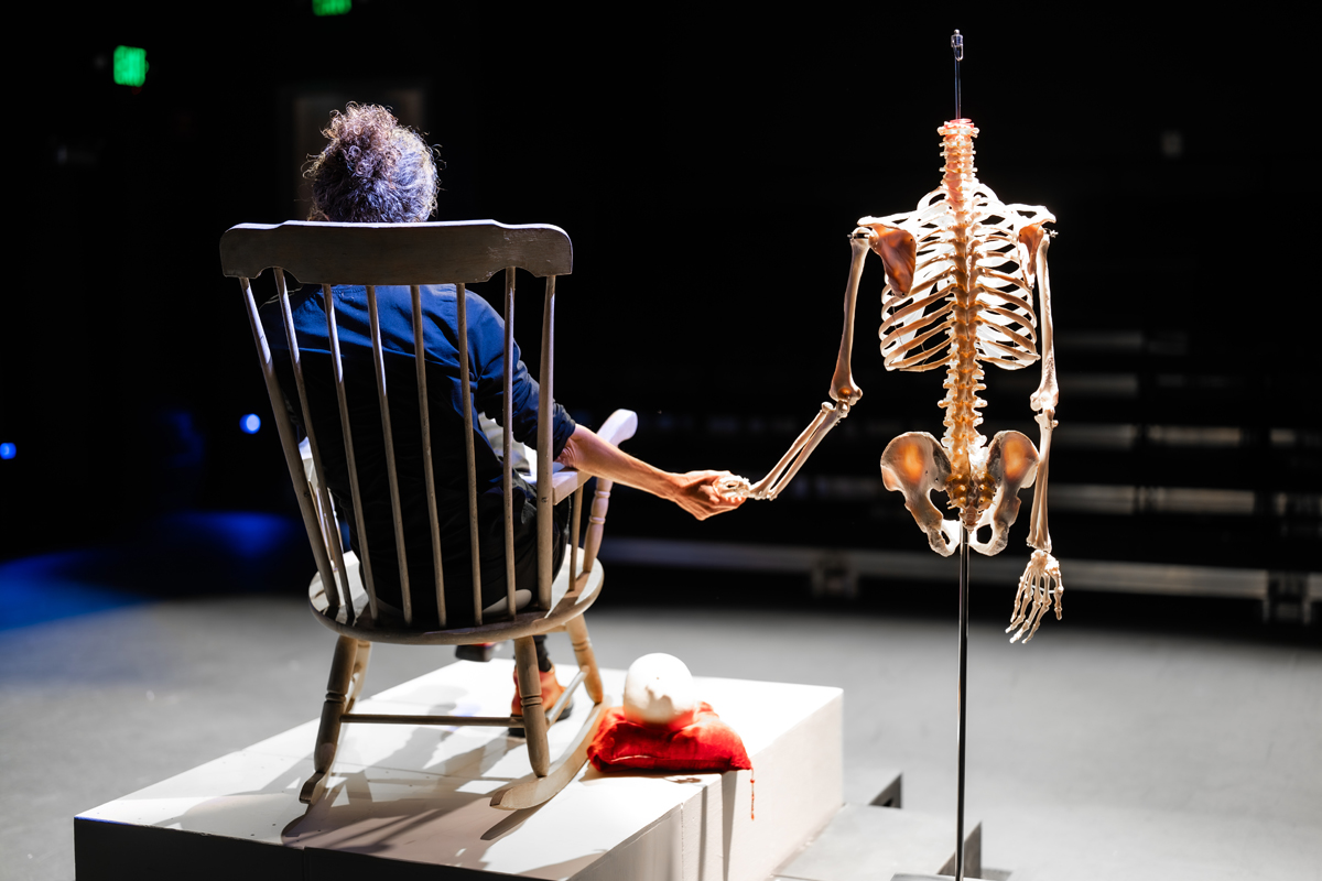 A seated woman holds hand with a skeleton during a production at Motus Theater, which has faced NEA grants cuts.