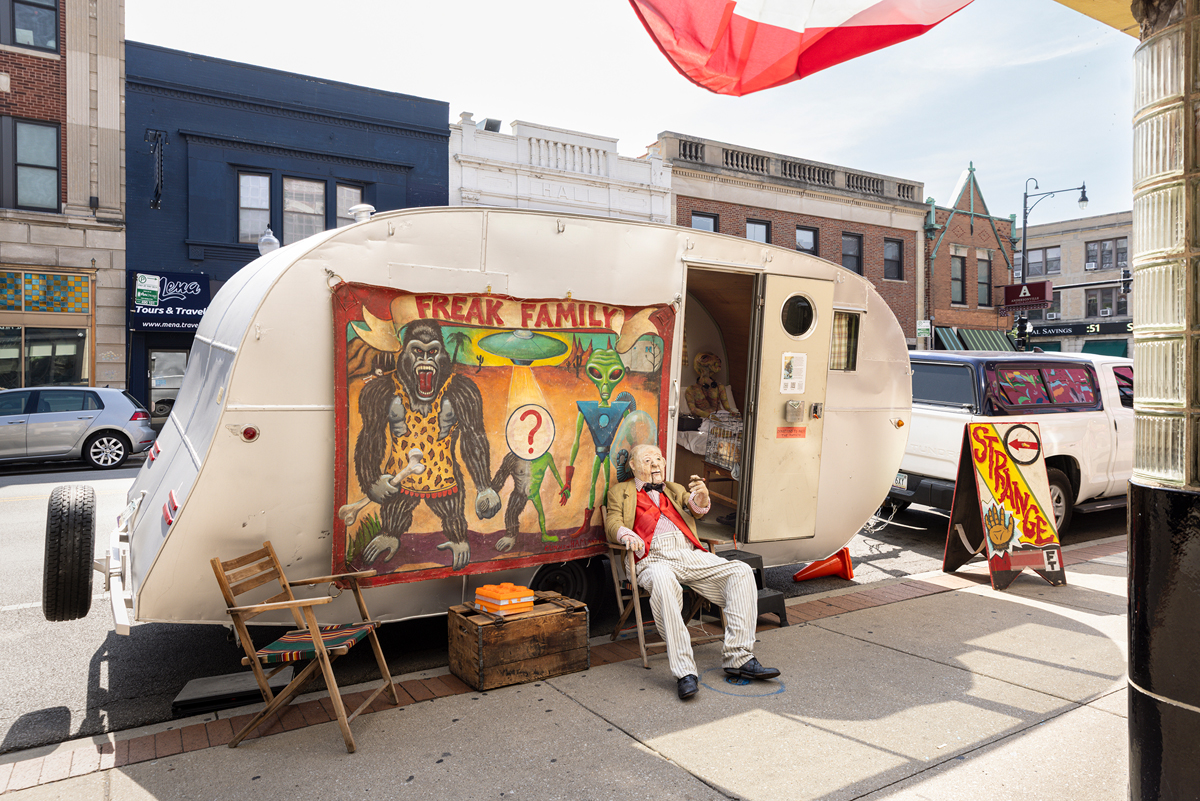 Trailer parked at a curb with roadshow exhibition inside and a mannequin sitting in a chair outside.