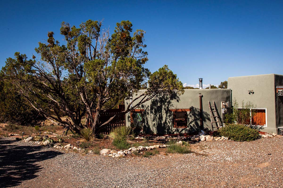 Image of the Aletheia House, an adobe casita and experimental creative space under the shade of a cedar/juniper tree in New Mexico.